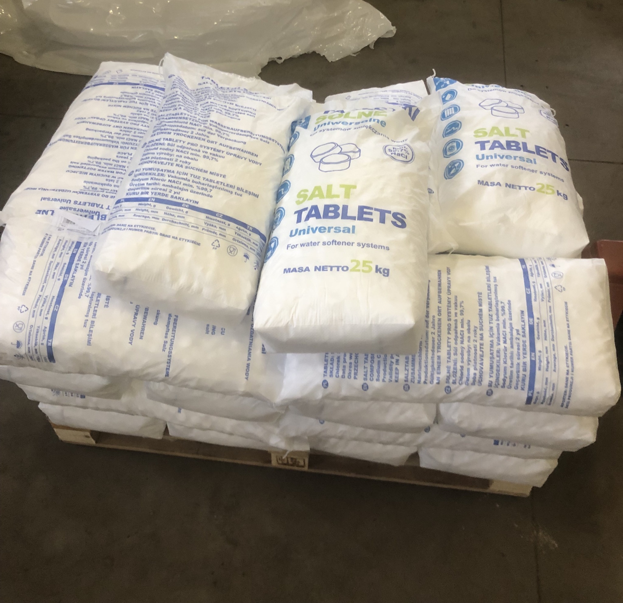 Paleta z workami białych tabletek solnych o wadze 25 kg, przeznaczonych do systemów uzdatniania wody, z widocznymi napisami w różnych językach.