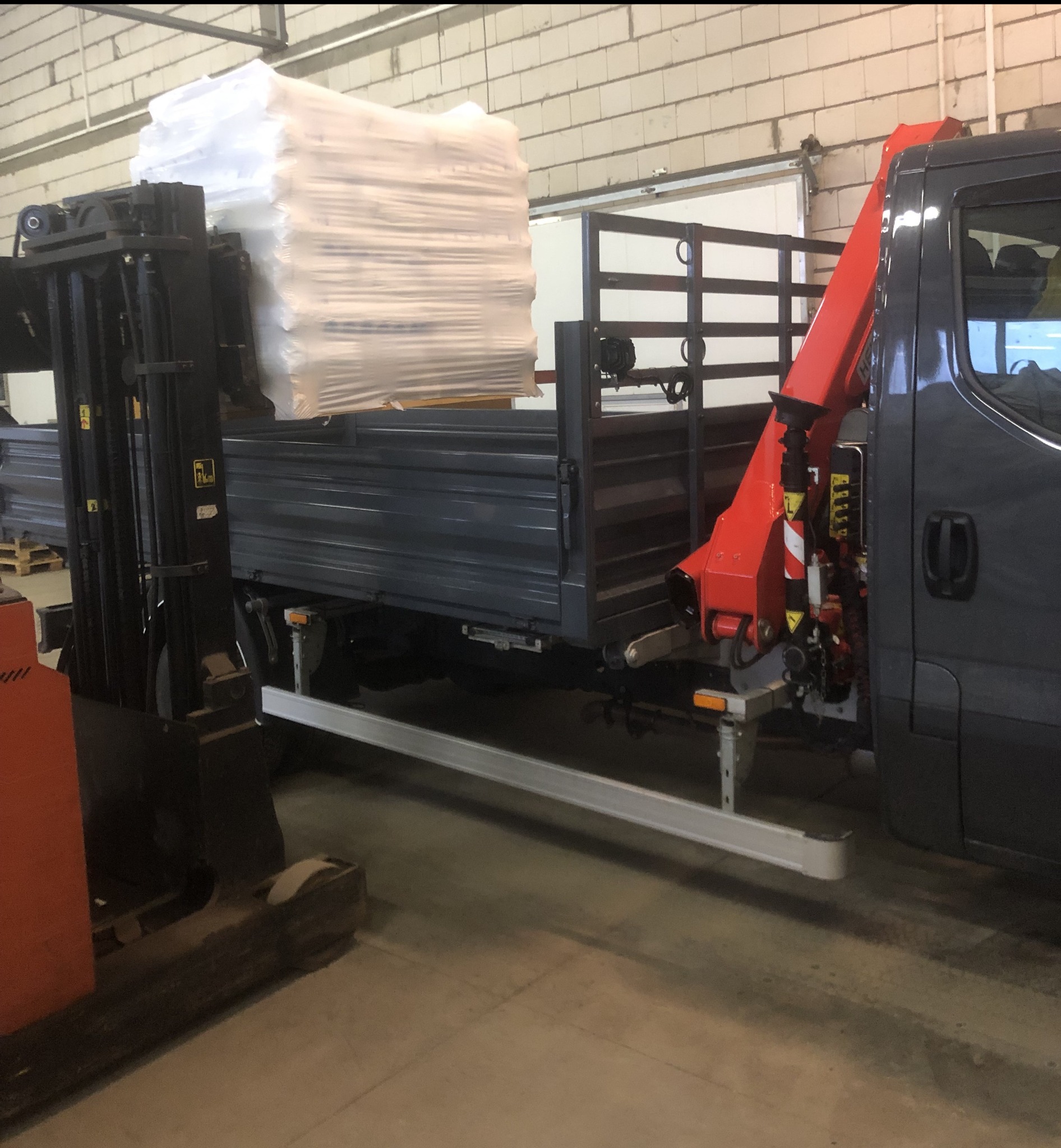 Wózek widłowy unosi paletę z towarem w folii, ładując ją na pakę samochodu z dźwigiem hydraulicznym w hali magazynowej.