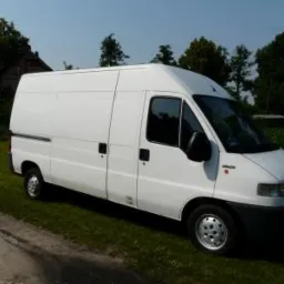 Biały, wysoki furgon marki Fiat Ducato, zaparkowany na trawie obok polnej drogi, w tle drzewa i budynek.