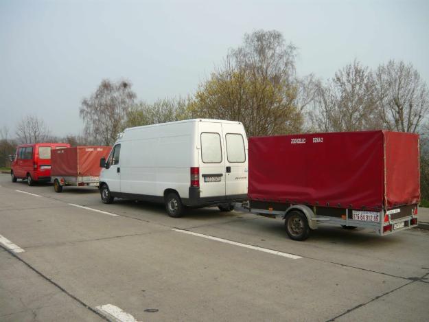 Biały bus z czerwoną plandeką przyczepy na parkingu w Dzierżoniowie, w tle drugi bus z przyczepą.