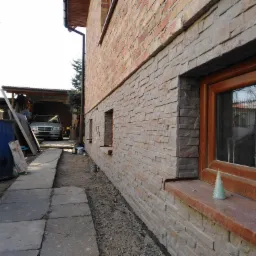 Elewacja domu z cegły i kamienia dekoracyjnego, widok z boku, z oknem i dekoracją w kształcie choinki na parapecie, w tle zaparkowany samochód.