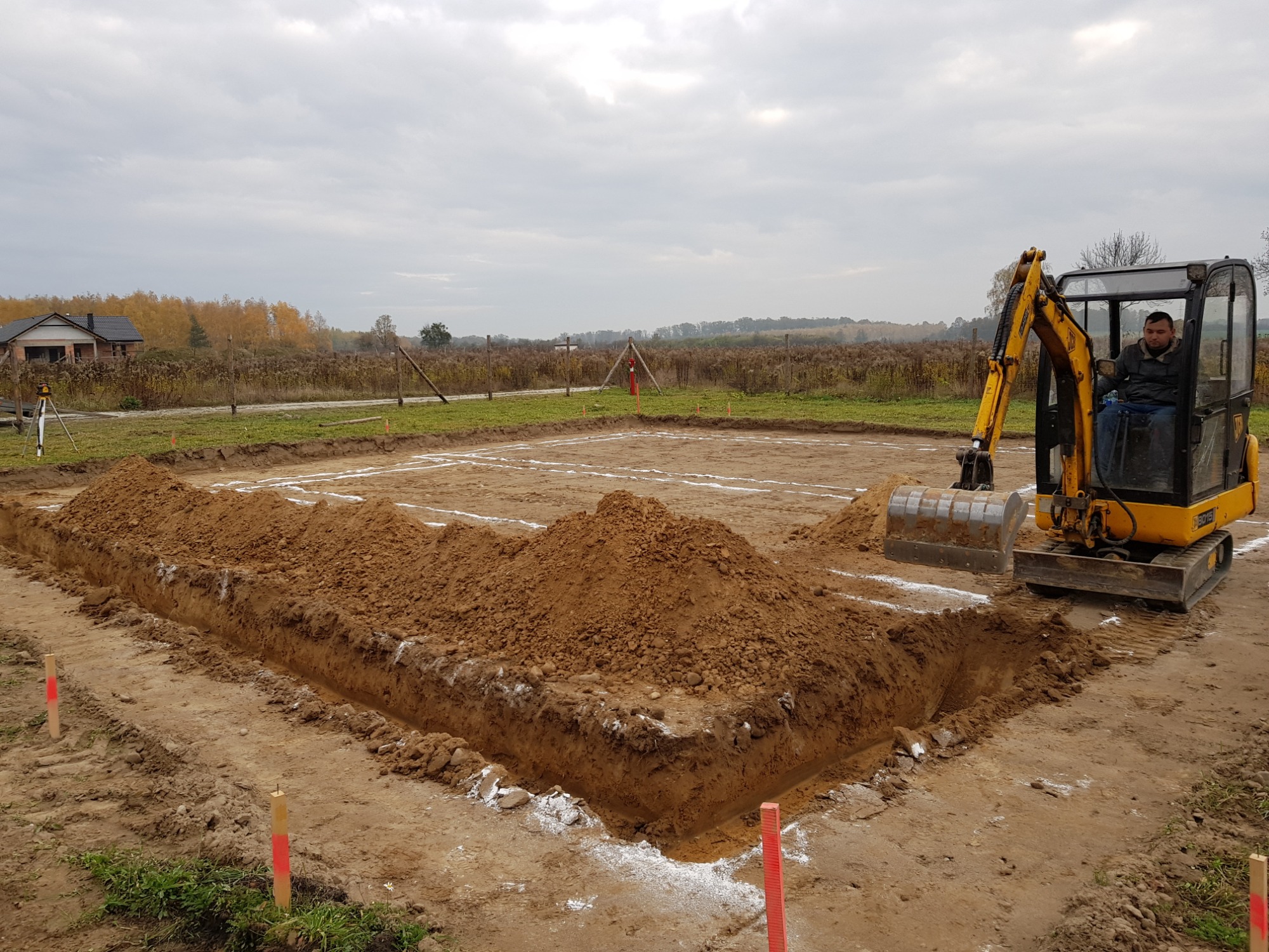 Żółta mini koparka z operatorem wykopuje rów pod fundamenty budynku, wyznaczone białym wapnem, w tle widoczny dom i jesienne drzewa.