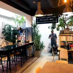 Wnętrze biura coworkingowego z wysokimi stołkami barowymi przy czarnym stole, rowerem opartym o ścianę, regałami z książkami i osobami pracującymi przy biurkach.