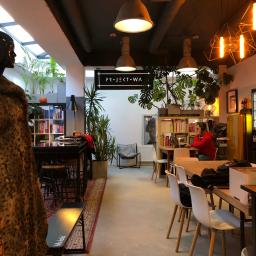 Projektowa Coworking Design Studio - Wnętrze kawiarni w stylu loft z lampami wiszącymi, roślinami doniczkowymi, stolikami z krzesłami, regałami z książkami i manekinem w futrze, widoczne logo 'Projekt WA'.