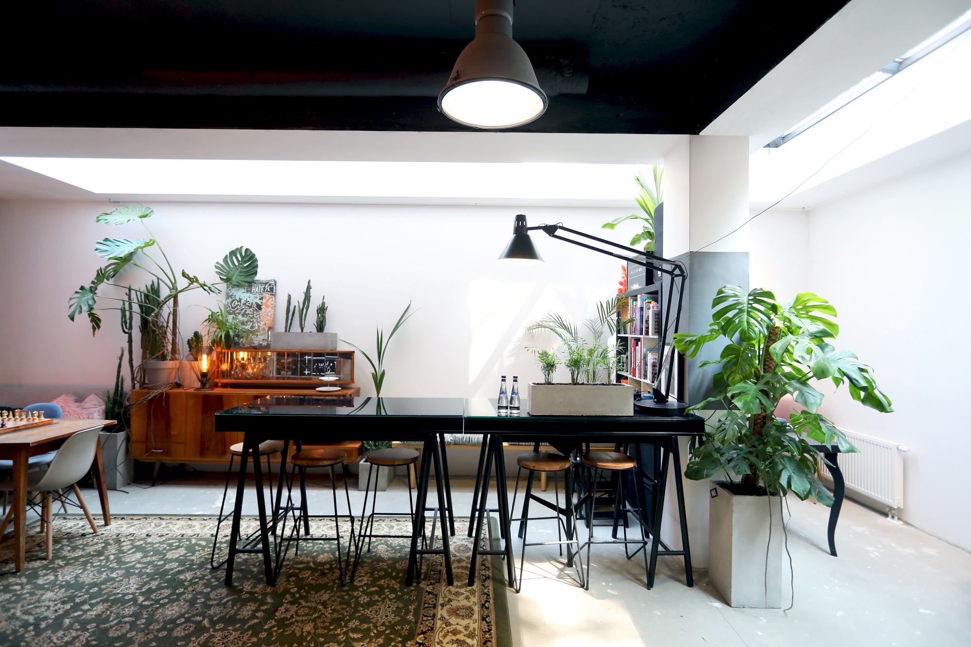 Jasne, przestronne wnętrze biura z wysokimi stołami barowymi, drewnianymi stołkami, roślinami doniczkowymi i vintage komodą w tle.