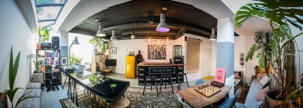 Wnętrze loftu z barem wykonanym z czarnych skrzynek, żółtą lodówką retro i stolikiem do szachów, oświetlone lampami przemysłowymi.