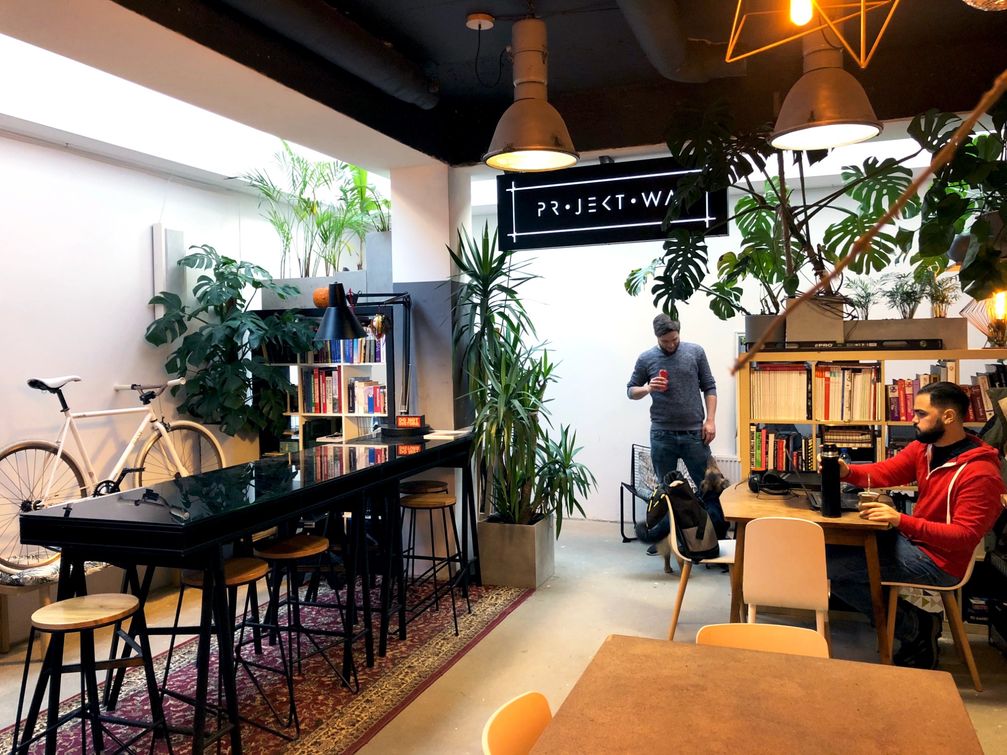 Wnętrze biura coworkingowego z wysokimi stołkami barowymi przy czarnym stole, rowerem opartym o ścianę, regałami z książkami i osobami pracującymi przy biurkach.