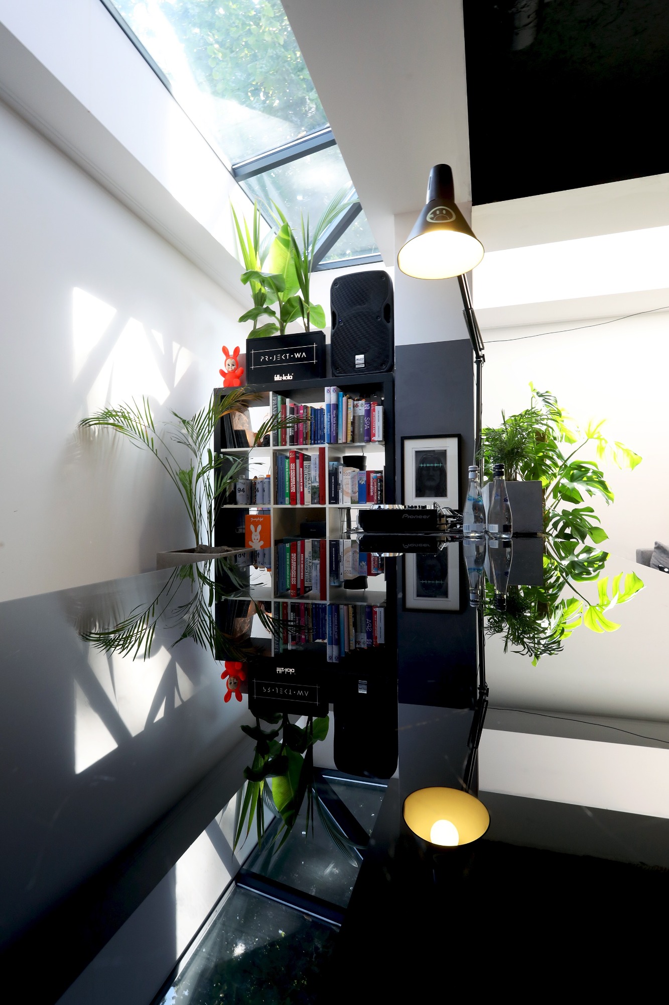 Nowoczesne wnętrze z czarnym, błyszczącym stołem, regałem z książkami, głośnikiem, roślinami doniczkowymi i lampą biurkową, oświetlone światłem wpadającym przez okno dachowe.