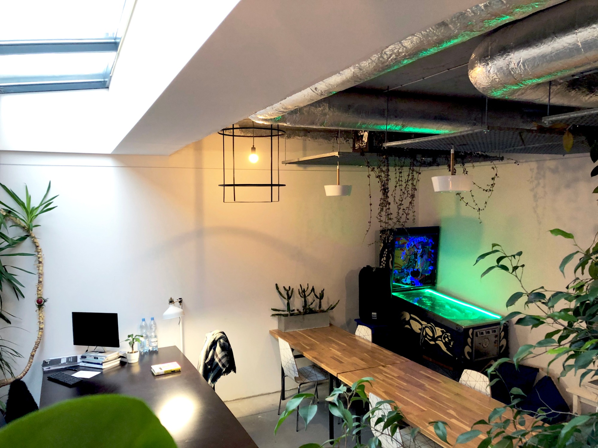 Jasne, przestronne biuro z drewnianym stołem, komputerem, roślinami doniczkowymi i automatem do gry w pinball z podświetleniem LED, oświetlone naturalnym światłem z okna dachowego.