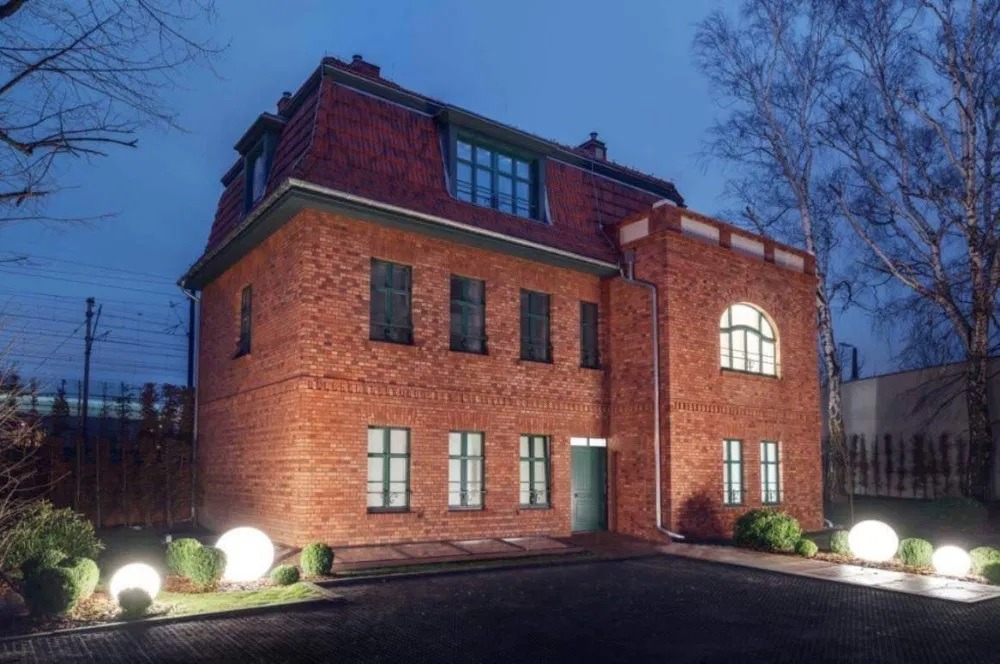 Dwupiętrowy budynek z czerwonej cegły z mansardowym dachem i zielonymi oknami, oświetlony kulistymi lampami ogrodowymi o zmierzchu.