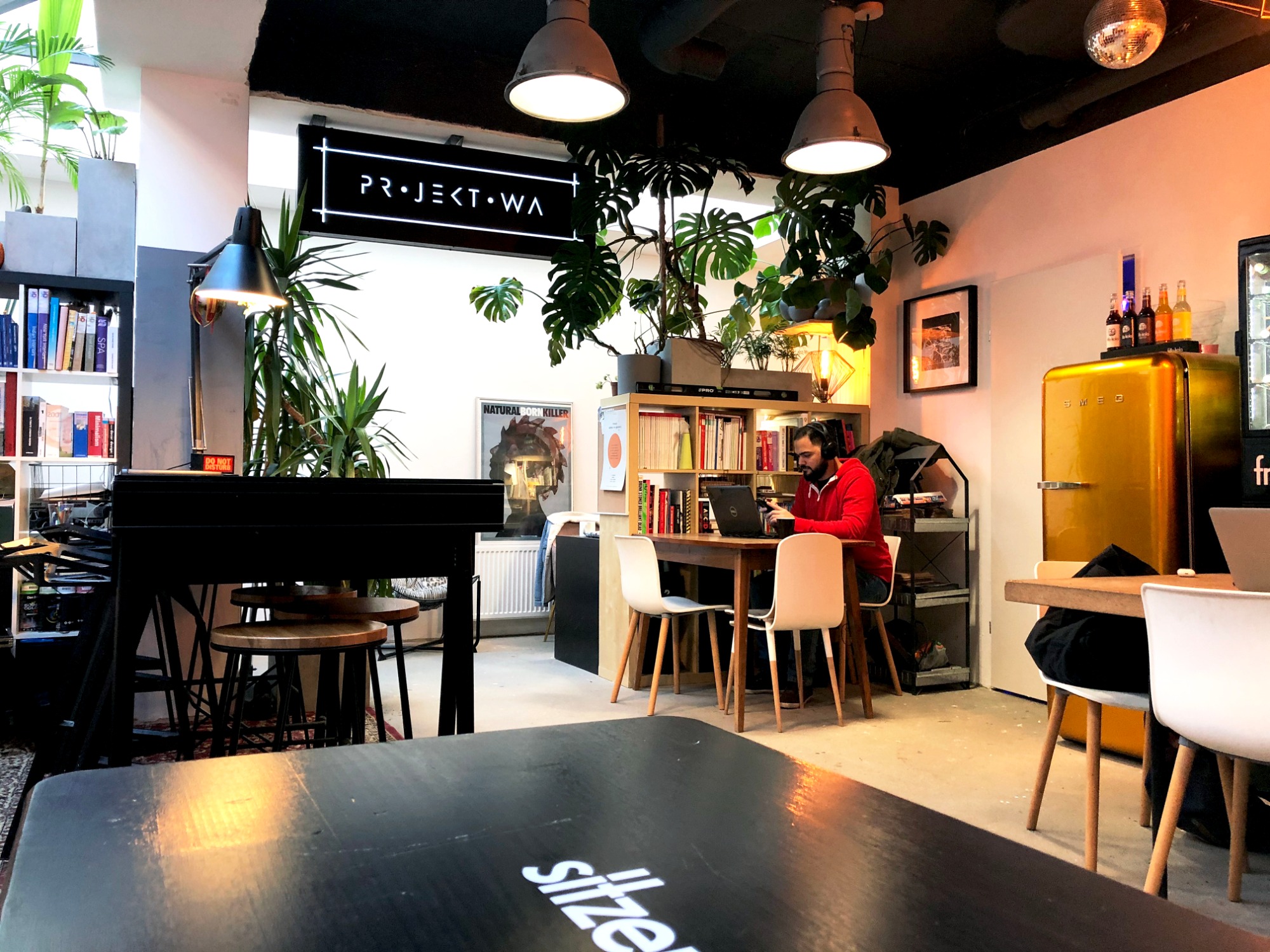 Wnętrze kawiarni lub biura coworkingowego z roślinami, lampami przemysłowymi, żółtą lodówką SMEG i osobą pracującą przy laptopie, widok z perspektywy stolika z logiem 'siltzer'.