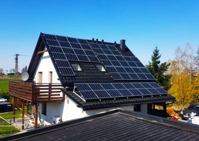 Dom jednorodzinny z czarną dachówką i panelami słonecznymi na dachu, balkon z drewnianą balustradą, antena satelitarna, błękitne niebo w tle.