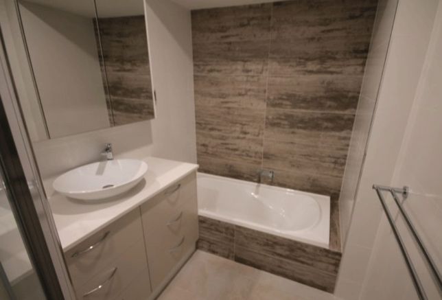 Nowoczesna łazienka z owalną umywalką na szafce z szufladami, wanną obudowaną płytkami imitującymi drewno i lustrem w szafce.