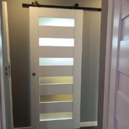 Białe przesuwne drzwi z matowego szkła, zawieszone na czarnym szynoprzewodzie, widoczne w otworze drzwiowym na tle szarej ściany i ciemnej podłogi.