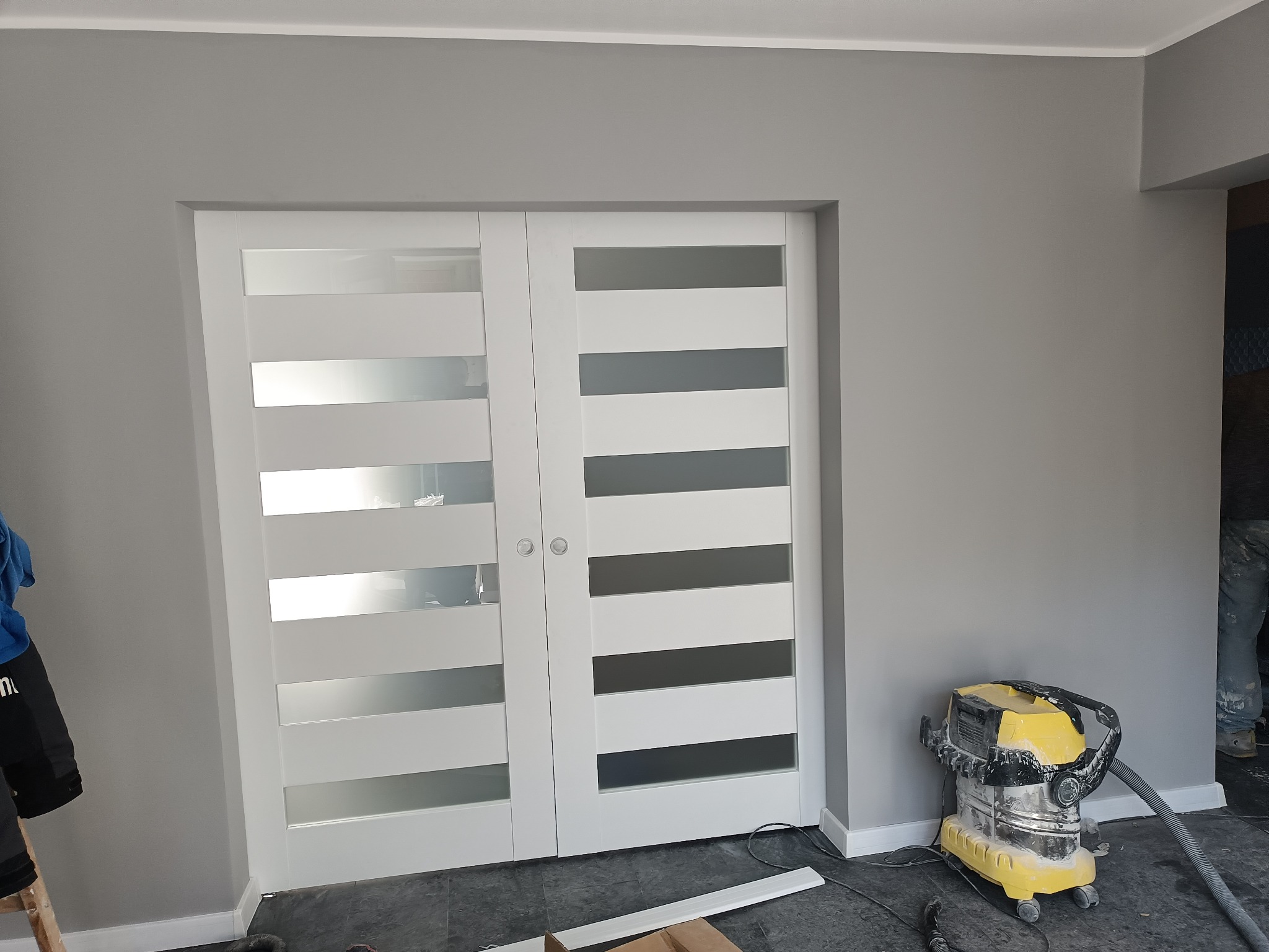 Nowoczesne białe drzwi przesuwne z poziomymi, przezroczystymi panelami na tle świeżo pomalowanej szarej ściany, widoczny odkurzacz przemysłowy i fragment drabiny.