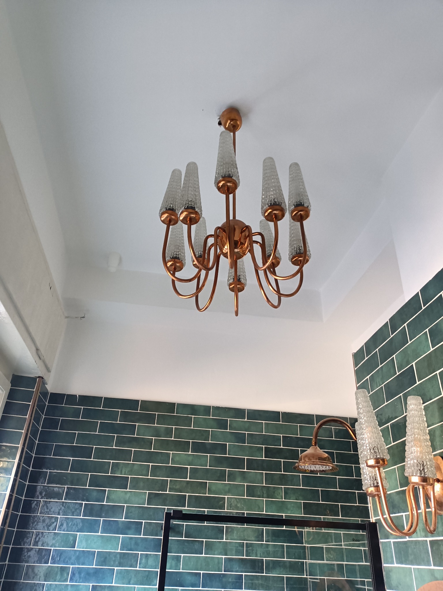 Miedziany żyrandol z kryształowymi kloszami oraz kinkiet nad kabiną prysznicową na tle ściany wyłożonej zielonymi, błyszczącymi płytkami imitującymi cegłę.