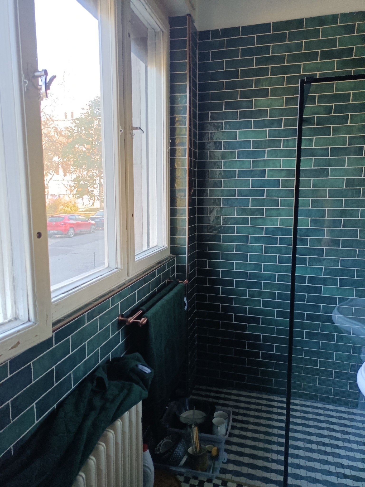 Wykończona łazienka z ciemnozielonymi, cegiełkowymi płytkami na ścianach, białą stolarką okienną z widokiem na ulicę, grzejnikiem z zawieszonym ręcznikiem i czarno-białą podłogą w zygzaki.