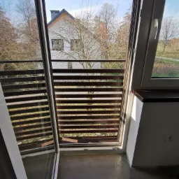 Widok z okna na balkon z poziomymi drewnianymi listwami, za nim dom jednorodzinny i drzewa w jesiennej scenerii.