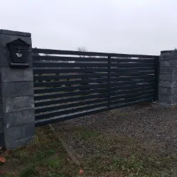 Nowoczesna, pozioma brama wjazdowa w kolorze antracytowym, z przesuwnymi panelami, osadzona na słupkach z szarej kostki łupanej, z czarną skrzynką na listy po lewej stronie, na tle szarego nieba...