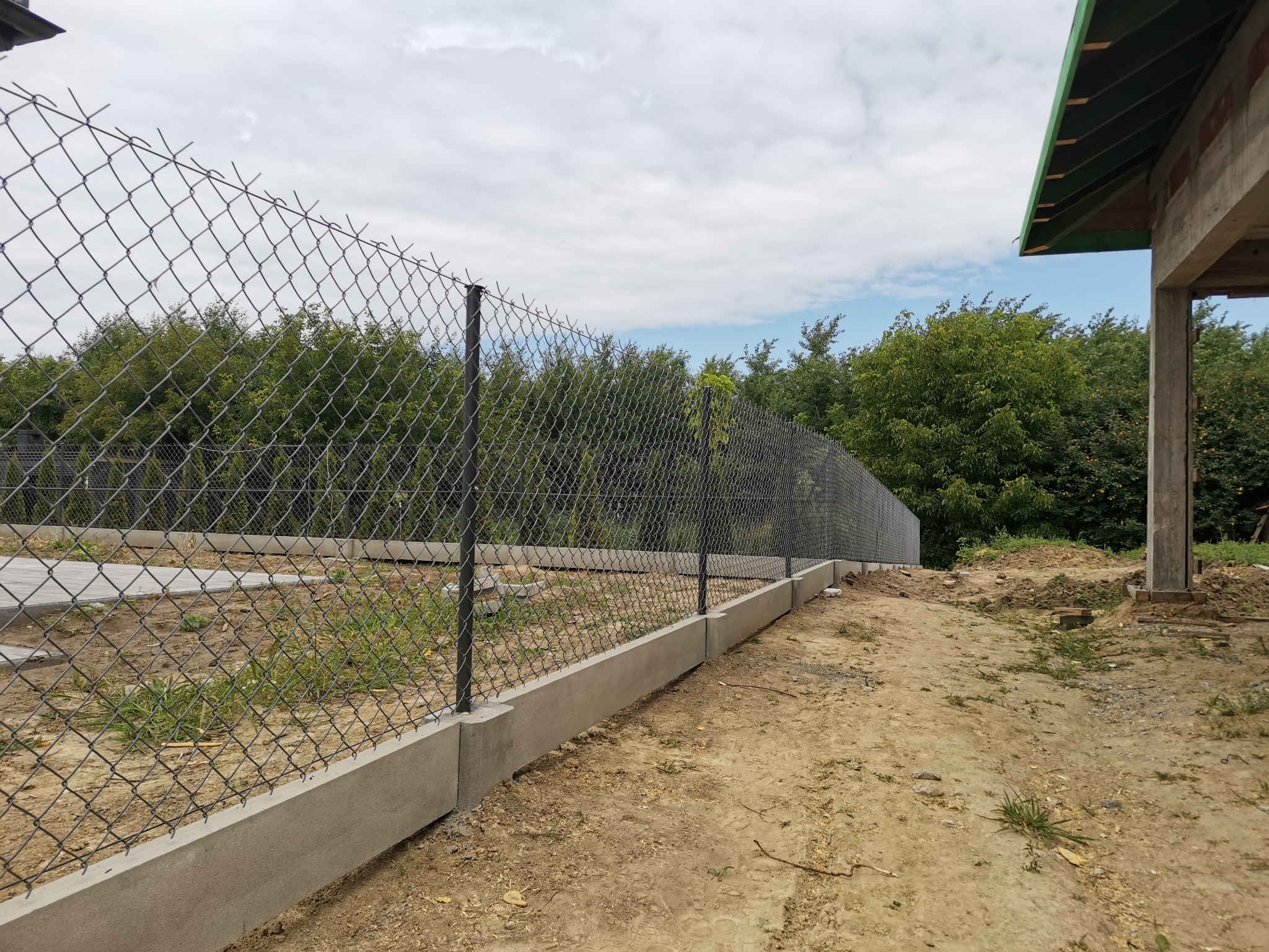 Długa, czarna siatka ogrodzeniowa z betonową podmurówką, widok z boku na tle drzew i częściowo wykończonego budynku w surowym stanie.