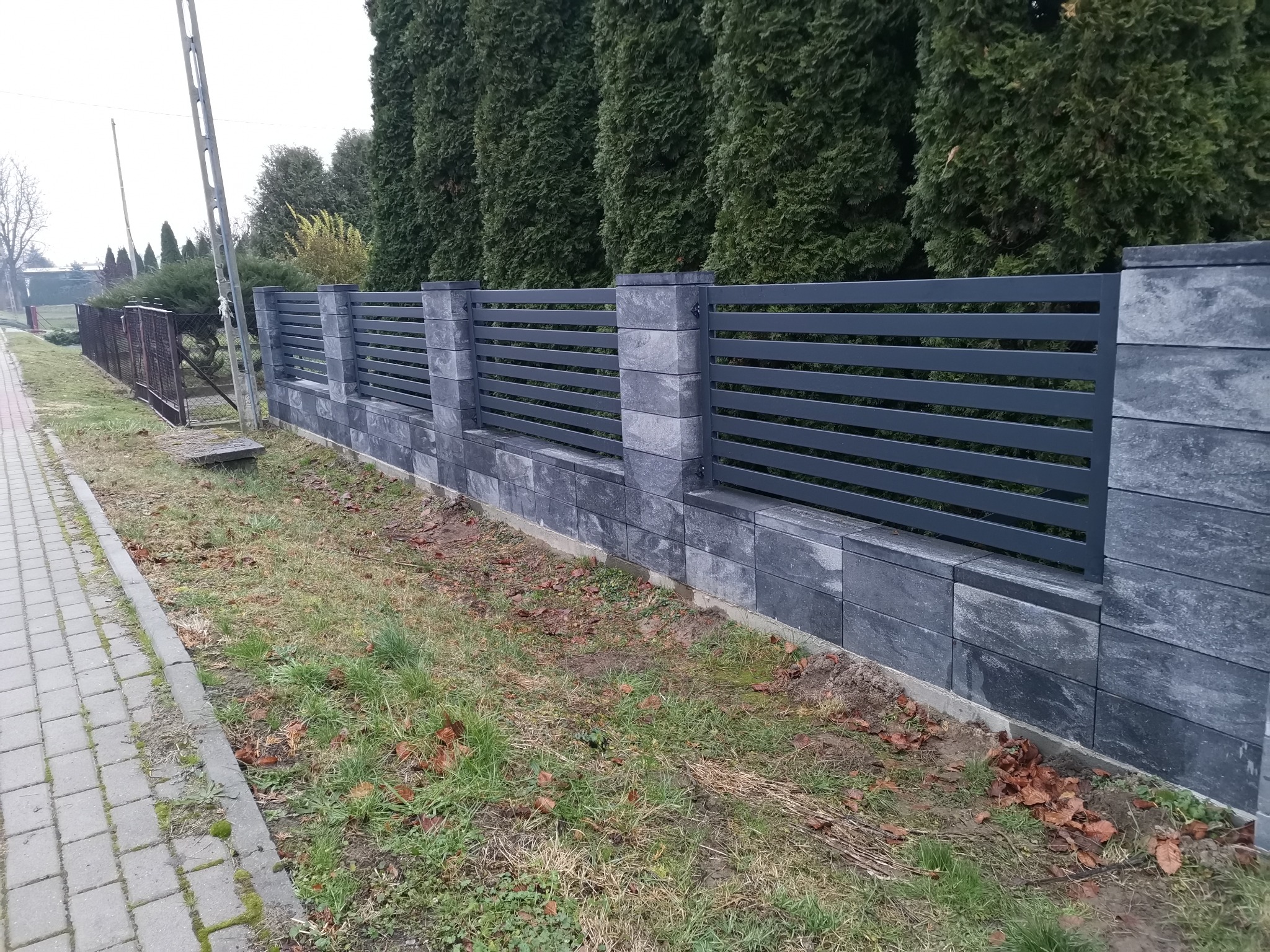 Nowoczesne ogrodzenie z poziomych, szarych paneli metalowych osadzonych na murowanych słupkach z szarej kostki, tło stanowią wysokie tuje.