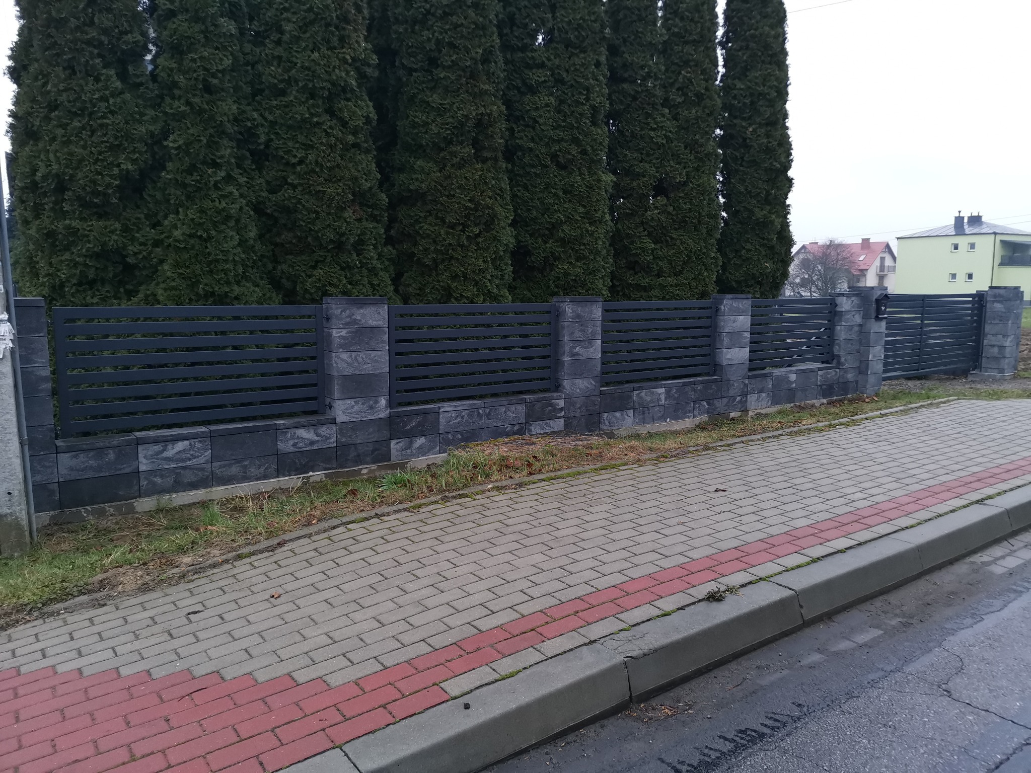 Nowoczesne ogrodzenie panelowe z poziomymi przęsłami w kolorze antracytowym, wsparte na słupkach z szarej kostki, na tle rzędu wysokich, zielonych tuj.