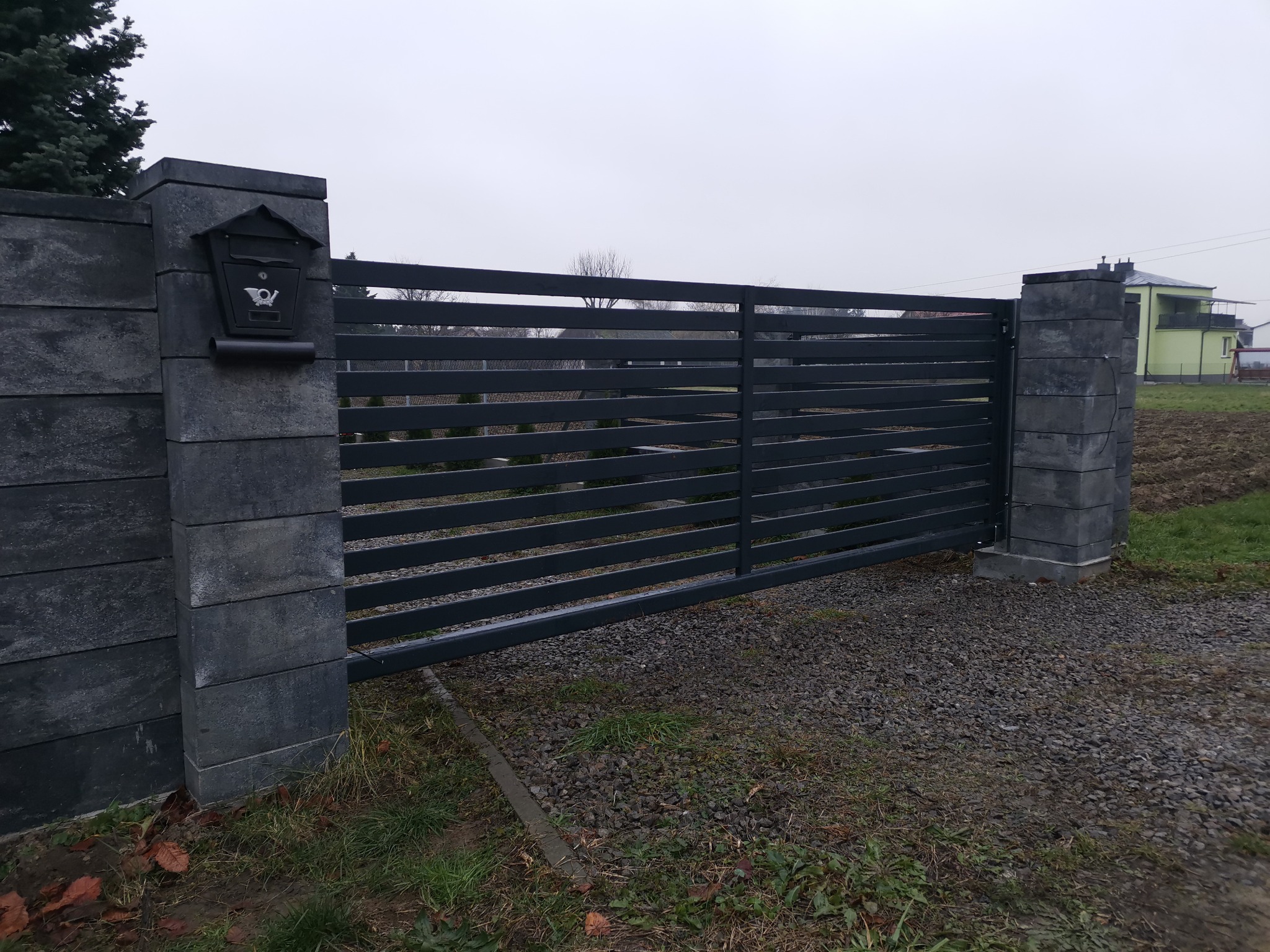 Nowoczesna, pozioma brama wjazdowa w kolorze antracytowym, z przesuwnymi panelami, osadzona na słupkach z szarej kostki łupanej, z czarną skrzynką na listy po lewej stronie, na tle szarego nieba...