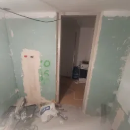 Wnętrze w trakcie prac remontowych: ściany z widocznymi śladami szpachlowania i zielonymi napisami, otwór drzwiowy, w tle widoczna kuchnia z kuchenką i butlą gazową, na podłodze narzędzia i worek...