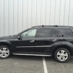 Czarny SUV Mercedes z relingami dachowymi, zaparkowany na parkingu z metalową ścianą w tle.