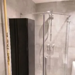 Nowoczesna kabina prysznicowa ze szklanymi drzwiami i chromowaną armaturą, obok czarna szafka łazienkowa i dekoracyjny bambusowy kij, szare płytki na ścianach.