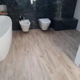 Nowoczesna łazienka z marmurową ścianą w odcieniach zieleni i beżową podłogą imitującą drewno, wolnostojąca wanna, bidet i toaleta w minimalistycznym stylu, oświetlona światłem wpadającym przez...