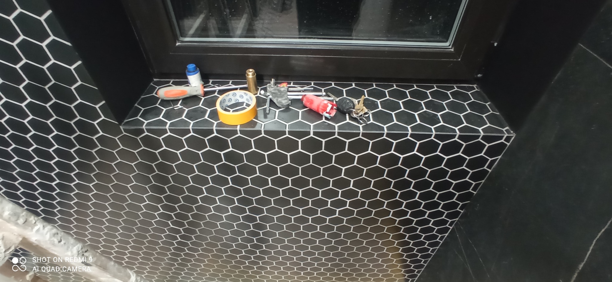 Ukończona ściana i parapet z czarnych, heksagonalnych płytek z białymi fugami. Na parapecie leżą narzędzia i akcesoria glazurnicze. Widoczne okno z czarną ramą.
