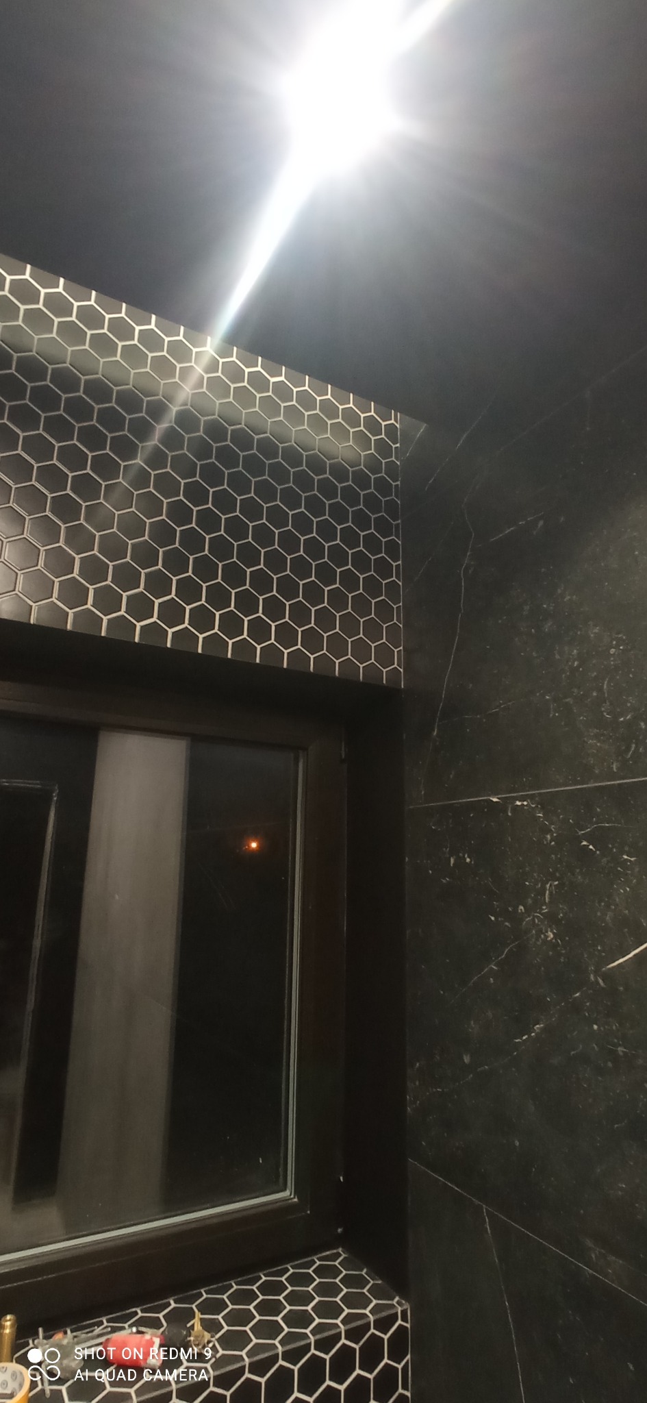 Częściowe ujęcie ściany z czarnych, heksagonalnych płytek i marmurowej ściany w łazience, z fragmentem okna. Widoczne narzędzia glazurnicze.
