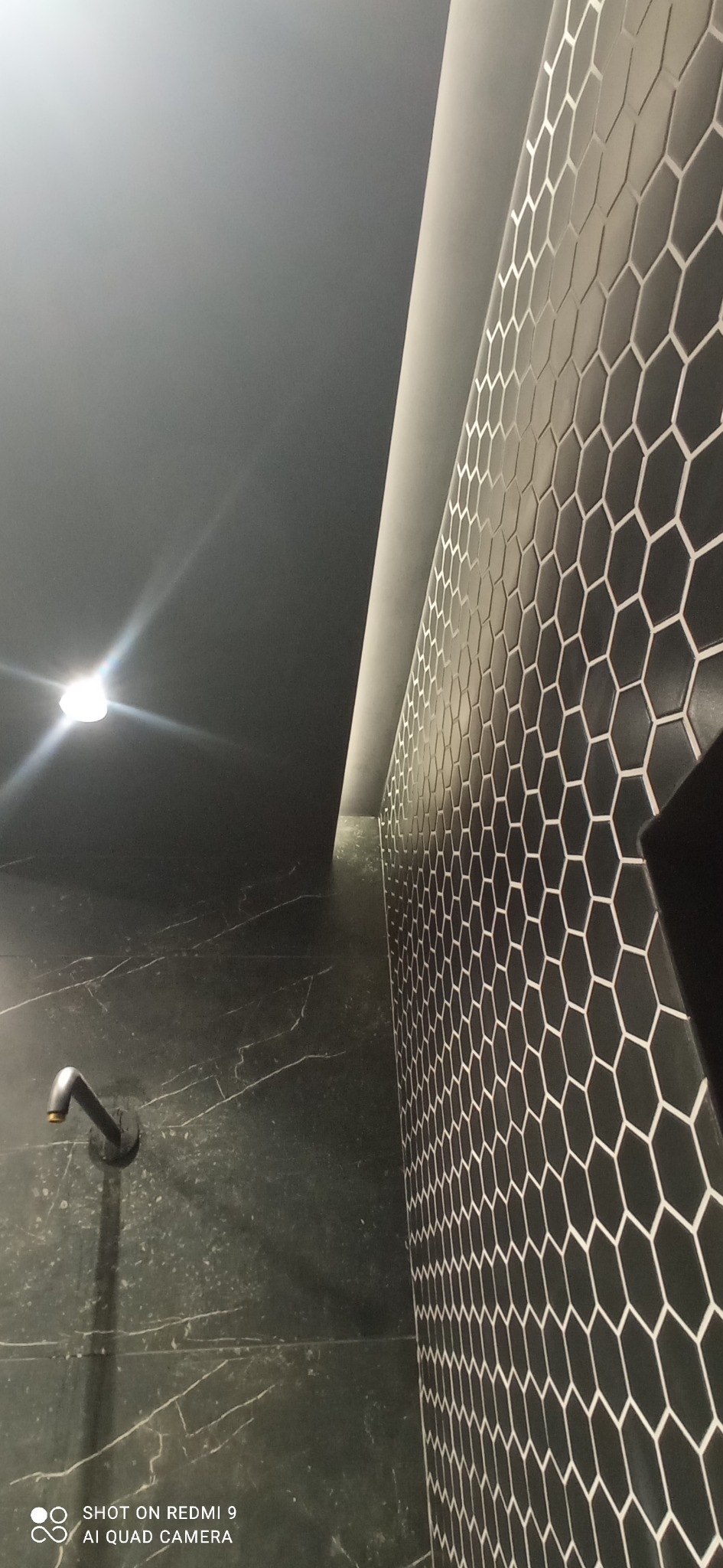 Nowoczesna łazienka z czarnymi heksagonalnymi płytkami na ścianie, marmurową ścianą i minimalistyczną armaturą. Oświetlenie LED podkreśla geometrię.