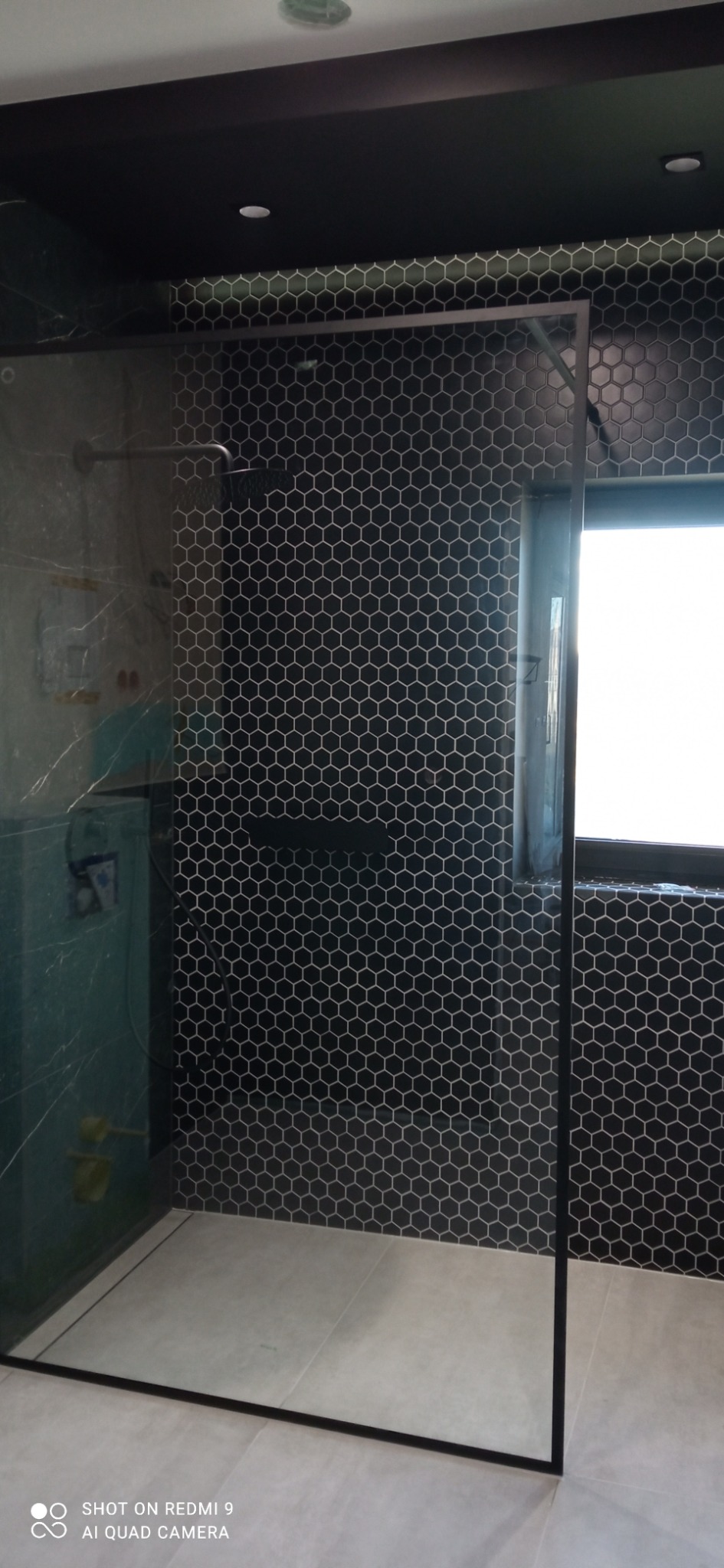 Nowoczesna kabina prysznicowa walk-in z czarnymi, heksagonalnymi płytkami na ścianach i szklaną ścianką z czarną ramą. Minimalistyczny design, jasne płytki podłogowe.