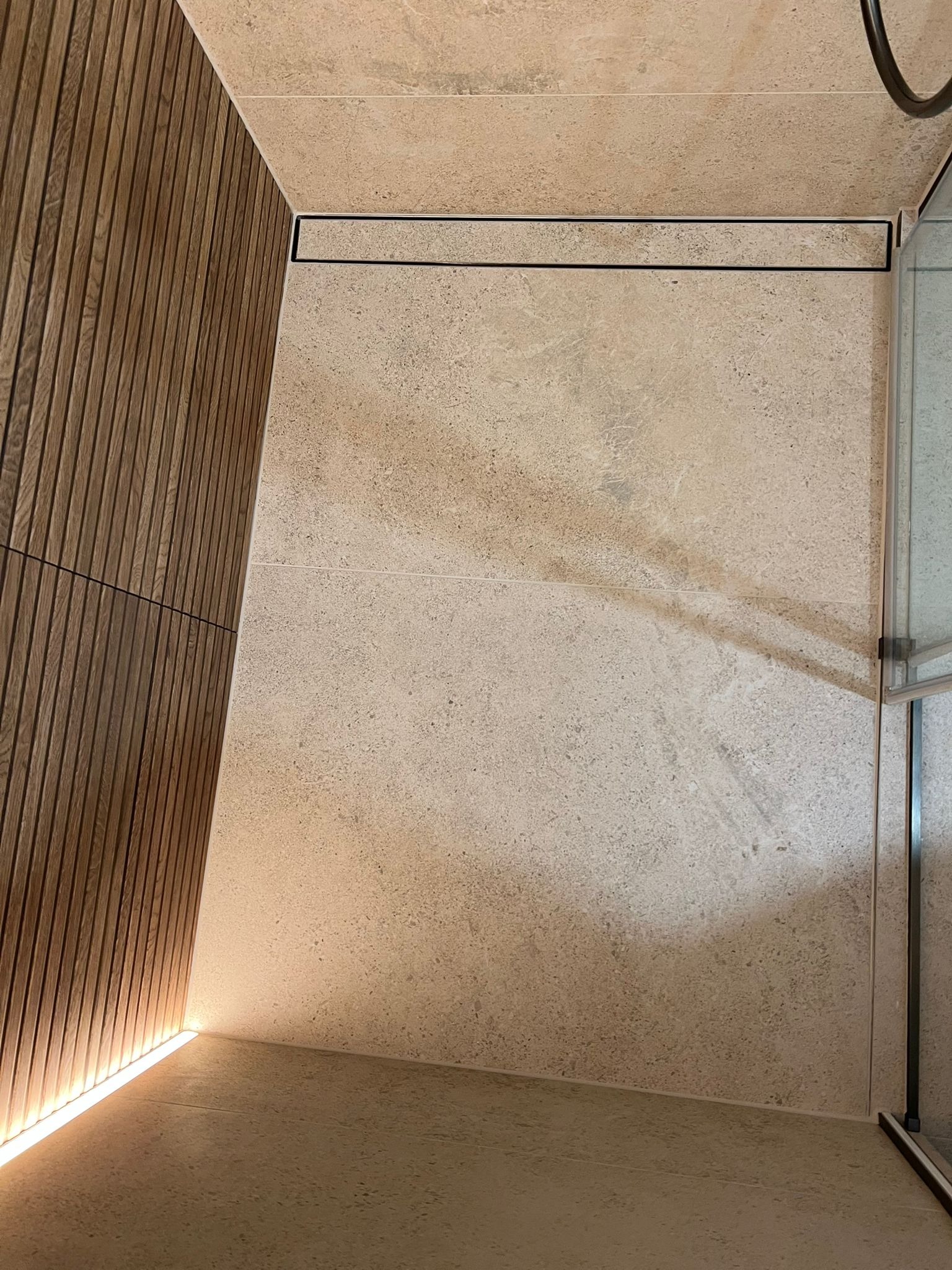 Nowoczesna łazienka z drewnianą ścianą i beżowymi płytkami. Minimalistyczny odpływ liniowy i subtelne oświetlenie LED podkreślają elegancję wykończenia.