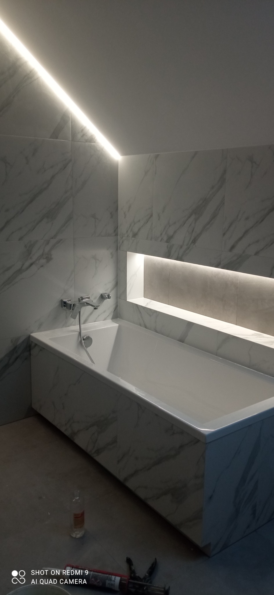 Wykończona łazienka z wanną obudowaną płytkami imitującymi marmur, podświetleniem LED na suficie i wnęką w ścianie. Widoczne narzędzia glazurnicze.