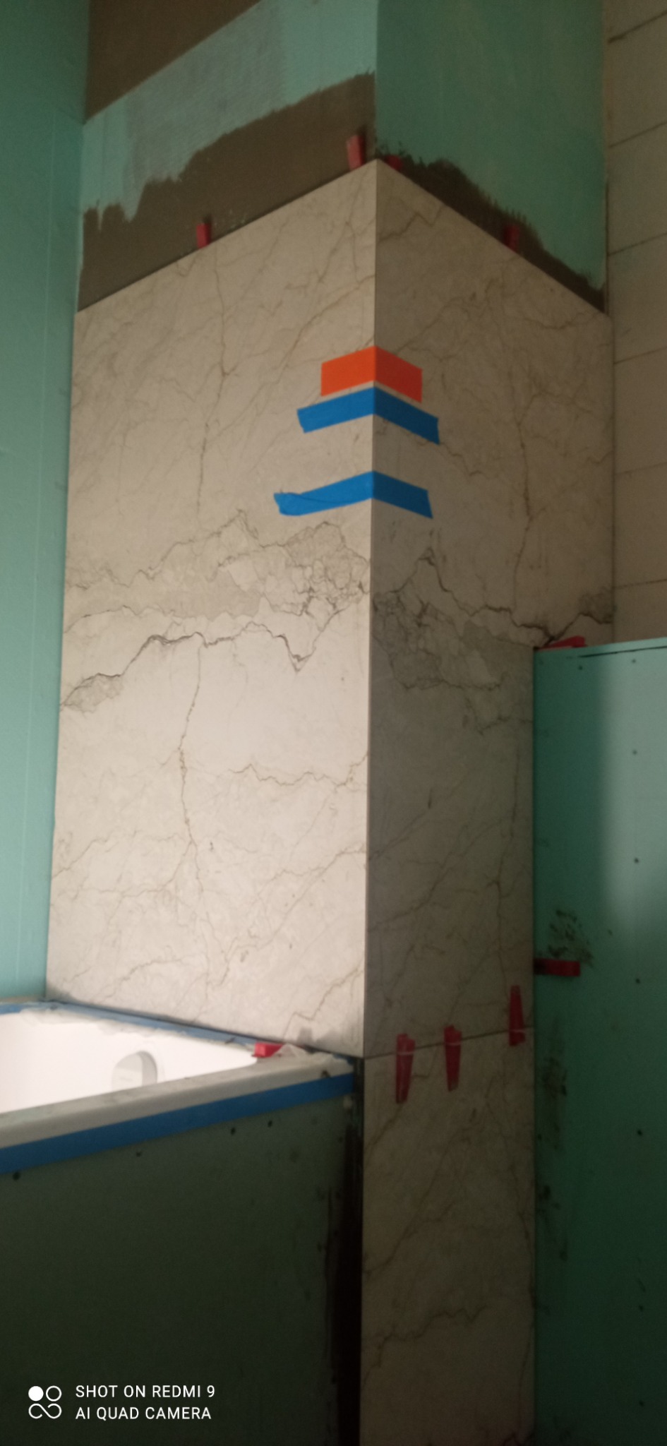 Fragment łazienki w trakcie prac glazurniczych: marmurowe płytki, poziomowanie z użyciem klipsów, niebieska taśma malarska, widoczna wanna i płyta GK.