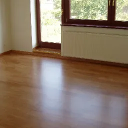 Jasne, lśniące panele podłogowe w pokoju z białymi ścianami, grzejnikiem pod oknem i widokiem na zieleń za oknem.