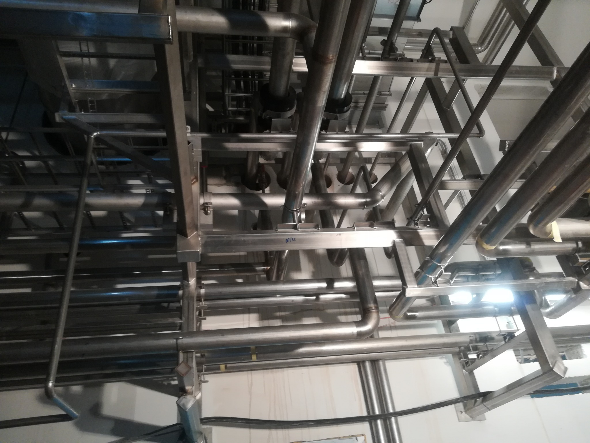 Kompleksowy system rur ze stali nierdzewnej, widok od dołu, tworzący skomplikowaną sieć w przemysłowym otoczeniu z jasnym oświetleniem i widocznymi spawami.