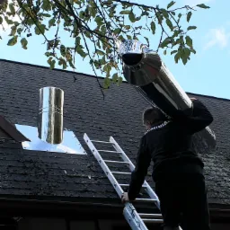 Pracownik w czarnej bluzie z napisem 'KOMINIAR' wchodzi po drabinie na dach z zamontowanym, nowym, błyszczącym, metalowym kominem, niosąc na ramieniu kolejny element komina.