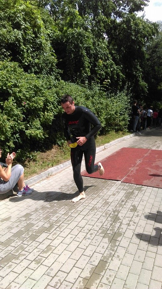 Mężczyzna w czarnym kombinezonie triathlonowym marki ZOOT biegnie boso po mokrej kostce brukowej, trzymając żółty czepek pływacki w dłoni, na tle zieleni i obserwujących osób.