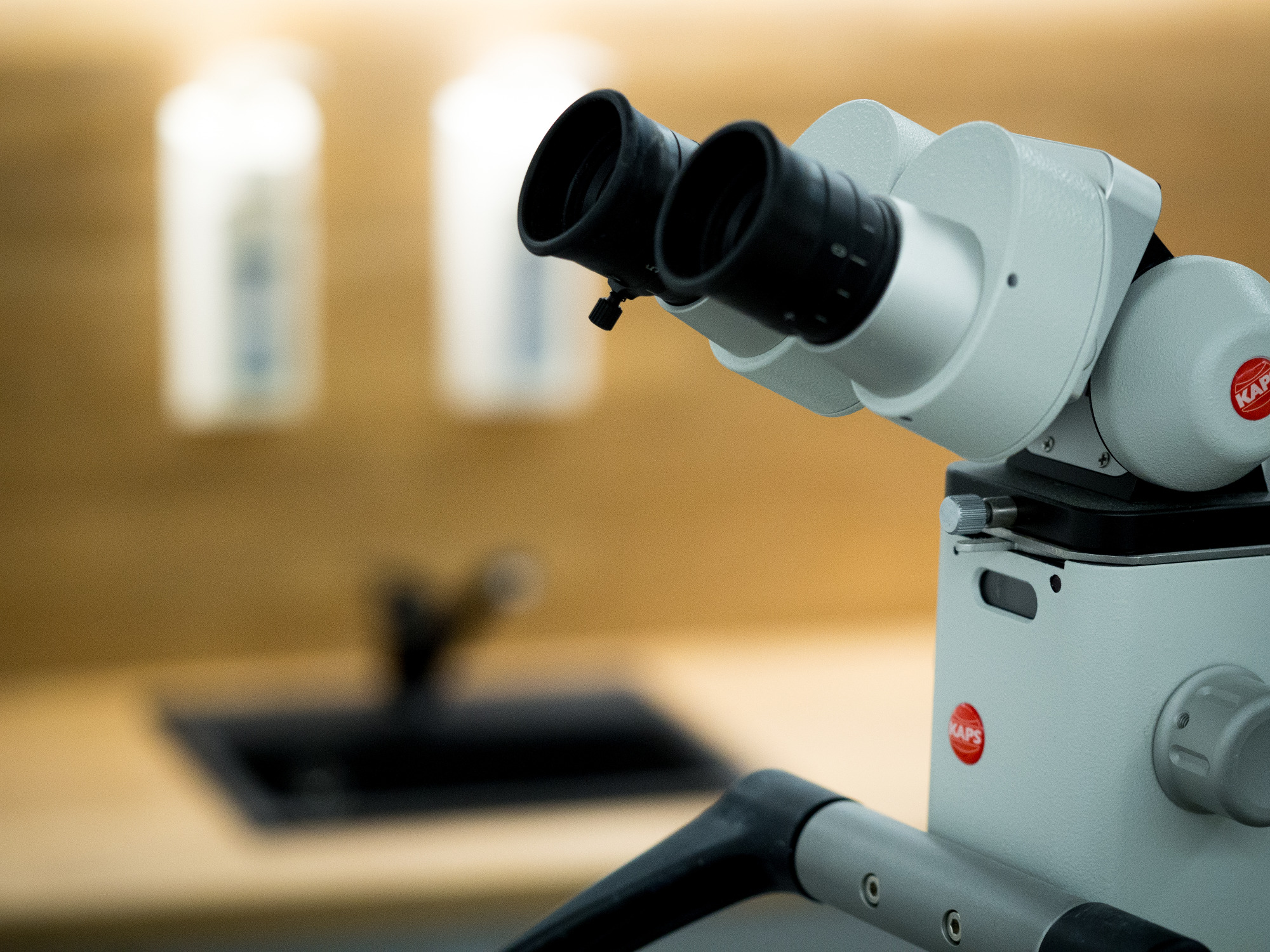 Stomatologiczny mikroskop operacyjny marki KAPS na tle blatu z umywalką i dozownikiem mydła w gabinecie.