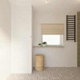 Jasna łazienka z wzorzystą podłogą, białą szafką z żaluzjowymi drzwiami, roletą rzymską w oknie, czarnym grzejnikiem drabinkowym i wiklinowym koszem na pranie.