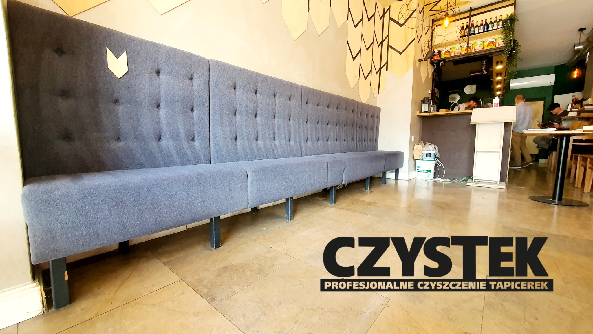 Długa, szara, tapicerowana ława z guzikami w restauracji, z logo firmy czyszczącej tapicerkę na pierwszym planie.