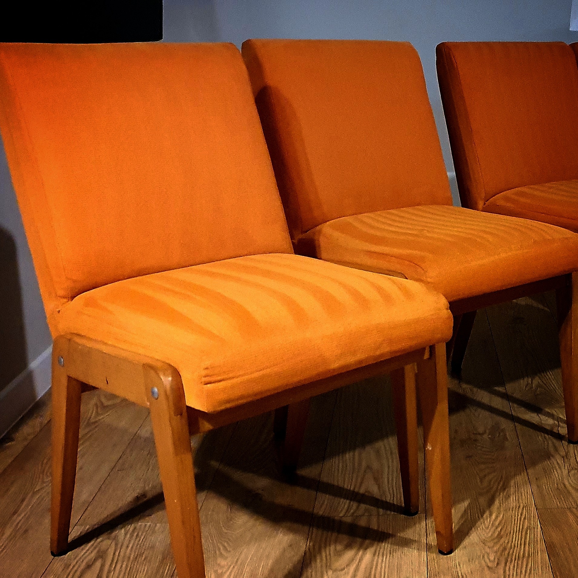 Trzy pomarańczowe krzesła tapicerowane z drewnianymi nogami ustawione rzędem na drewnianej podłodze, widoczne z bliska.