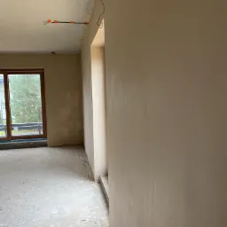 Wnętrze w stanie deweloperskim z oknem, surowym betonem na podłodze i świeżo położoną, jasną warstwą wykończeniową na ścianach, widoczne przewody elektryczne na suficie.