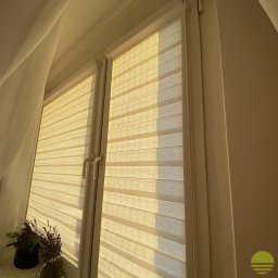 MARKIZA.PL SPÓŁKA Z OGRANICZONĄ ODPOWIEDZIALNOŚCIĄ - Beżowe, poziome pasy rolety okiennej dzień noc, częściowo zasłoniętej, z widocznym mechanizmem łańcuszkowym, w oknie PCV, z dekoracją roślinną na parapecie, oświetlone ciepłym światłem.