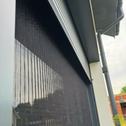 MARKIZA.PL SPÓŁKA Z OGRANICZONĄ ODPOWIEDZIALNOŚCIĄ - Czarna, zwijana przesłona okienna z widoczną strukturą siatki, zamontowana na elewacji budynku z rynną spustową. W tle zarys drzew i budowa.