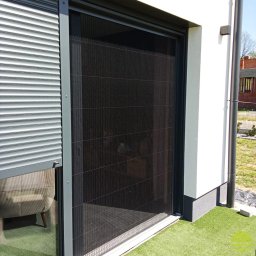 MARKIZA.PL SPÓŁKA Z OGRANICZONĄ ODPOWIEDZIALNOŚCIĄ - Zewnętrzna, plisowana moskitiera w kolorze czarnym zamontowana na oknie tarasowym, obok zwiniętej rolety zewnętrznej w kolorze srebrnym, na tle białej ściany budynku.